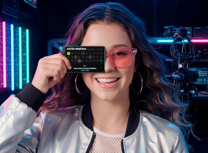 Uma artista sorrindo segura o Cartão Frequência da HZ Studios na frente do rosto, num studio com luzes neon